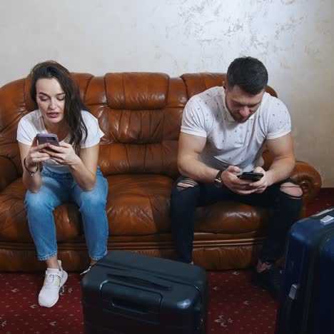 couple on couch with phones near suitcases. young woman and man waiting for the leaving from the hotel and playing on mobile phones