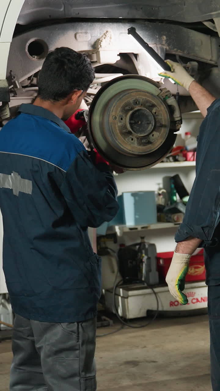 mecánico trabajando en la reparación de automóviles mientras los técnicos de laboratorio con tabletas observan y toman notas, taller mecánico con herramientas, vehículo levantado, colaboración e inspección técnica en progreso