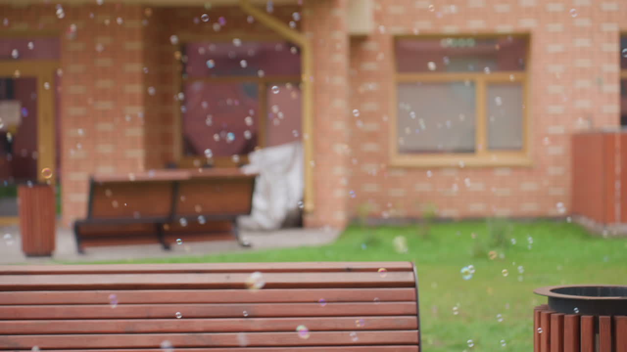 Floating soap bubbles drift across courtyard in front of brick building, wooden bench and green grass visible in soft blur, gentle airflow carries tiny spheres past windows