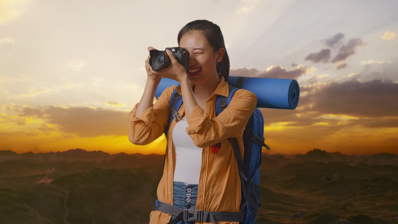 excursionista asiática con mochila de montañismo usando una cámara tomando fotos mientras está de pie en la cima de la montaña durante el atardecer