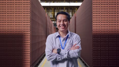 shot captures of an asian male professional worker standing with red brick packed in stacks are stored, his broad smile at the camera and cross his arm over his chest