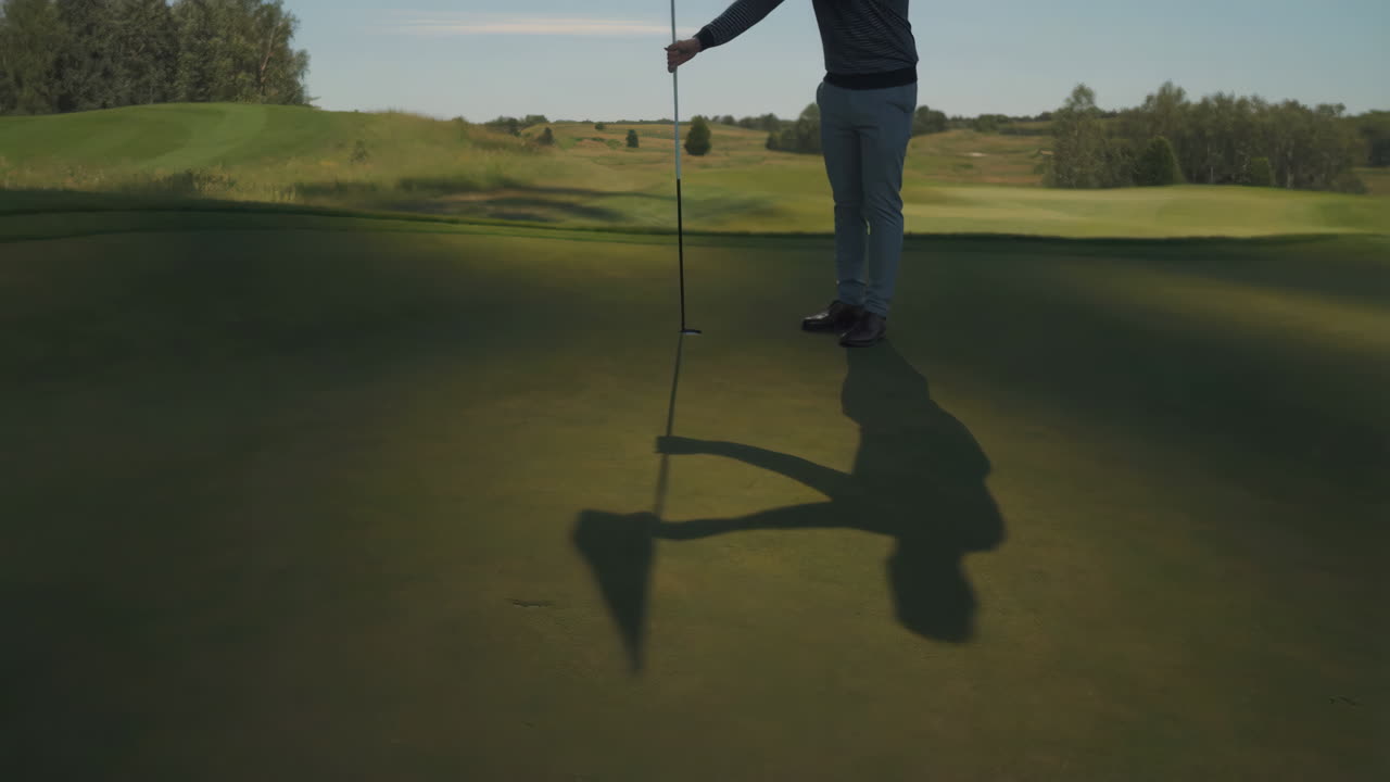 Golfista che tiene l'asta della bandiera su un putting green, proiettando una lunga ombra