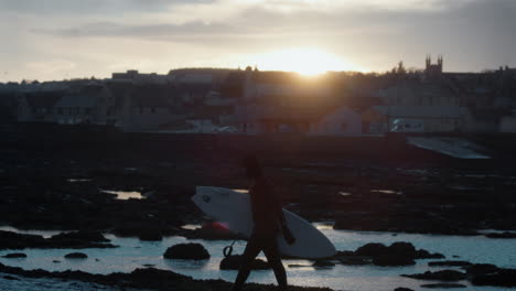 El-Sol-Poniente-Brilla-Sobre-Una-Ciudad-Costera-En-Irlanda-Mientras-Un-Surfista-En-Traje-De-Neopreno-Sale-Del-Agua-Bajo-Nubes-De-Tormenta