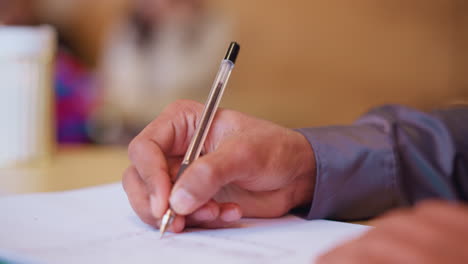 close-up hand holding pen while writing on white paper, focus on detailed grip and pen motion, person engaged in learning or office work, background softly blurred, calm indoor environment captured clearly