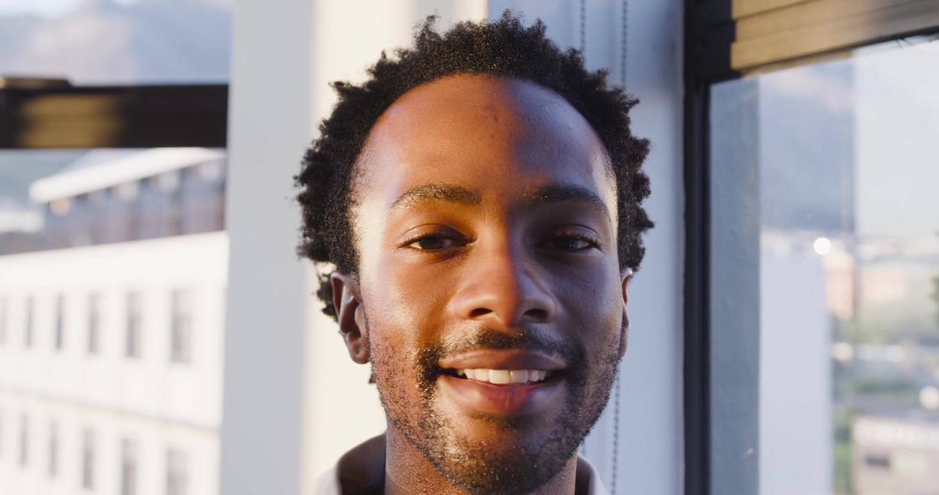 Adult African American male leaning at window sunlight falling turning to lens closing eyes smiling