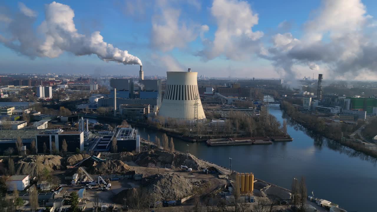 power plant emitting steam from its cooling tower, situated by a river and overlooking the cityscape. Smooth aerial view flight descending drone