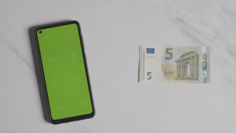 disparo de moneda aérea de la mano agarrando el billete de 5 euros junto a la pantalla verde del teléfono móvil