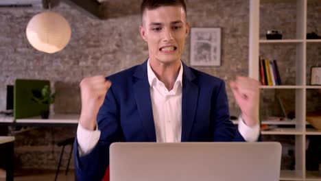young concentrated businessman is watching laptop in office, sees good results, keeping hand fist, yes gesture, business concept, communication concept
