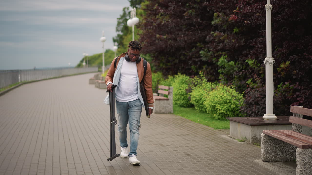 black man walking by waterfront, carrying DJ gear case, purposeful stride in casual jacket and jeans, urban promenade lined with benches and greenery, determined DJ and producer en route to outdoor