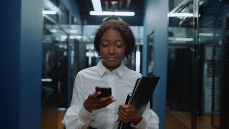 Afro-woman-having-phone-conversation-in-corridor.-African-lady-texting-in-office