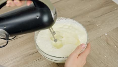 close-up top-down shot of electric mixer whipping egg yolk, mascarpone and powdered sugar in glass bowl on wood surface, creamy consistency forming, hand steadying bowl, real time, prep motion shot