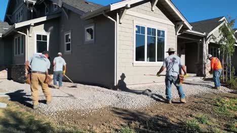 workers are busy laying gravel around a newly constructed home in a residential area. tools and teamwork are vital as they create a smooth surface by hand in bright sunlight.