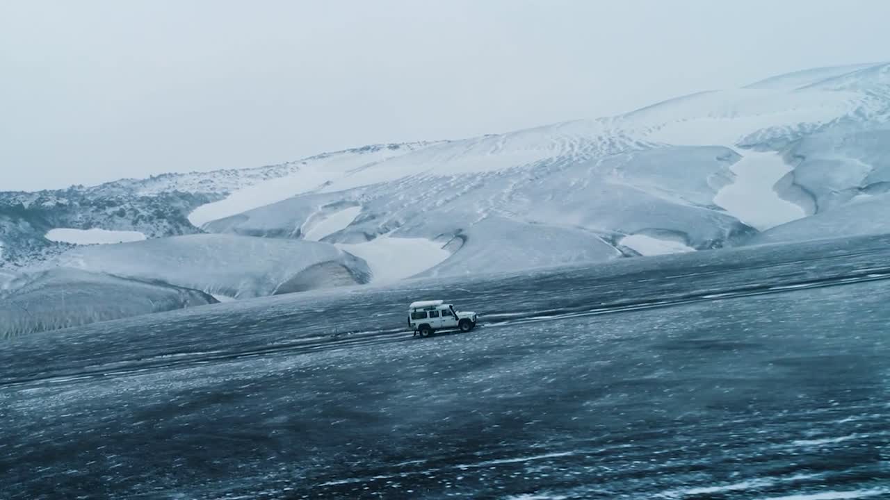 Aerial drone footage flying besides a white 4x4 vehicle truck while it is driving on a ice glacier or rural desolate road covered in ice and snow while mountains are in back at Greenland or Iceland.