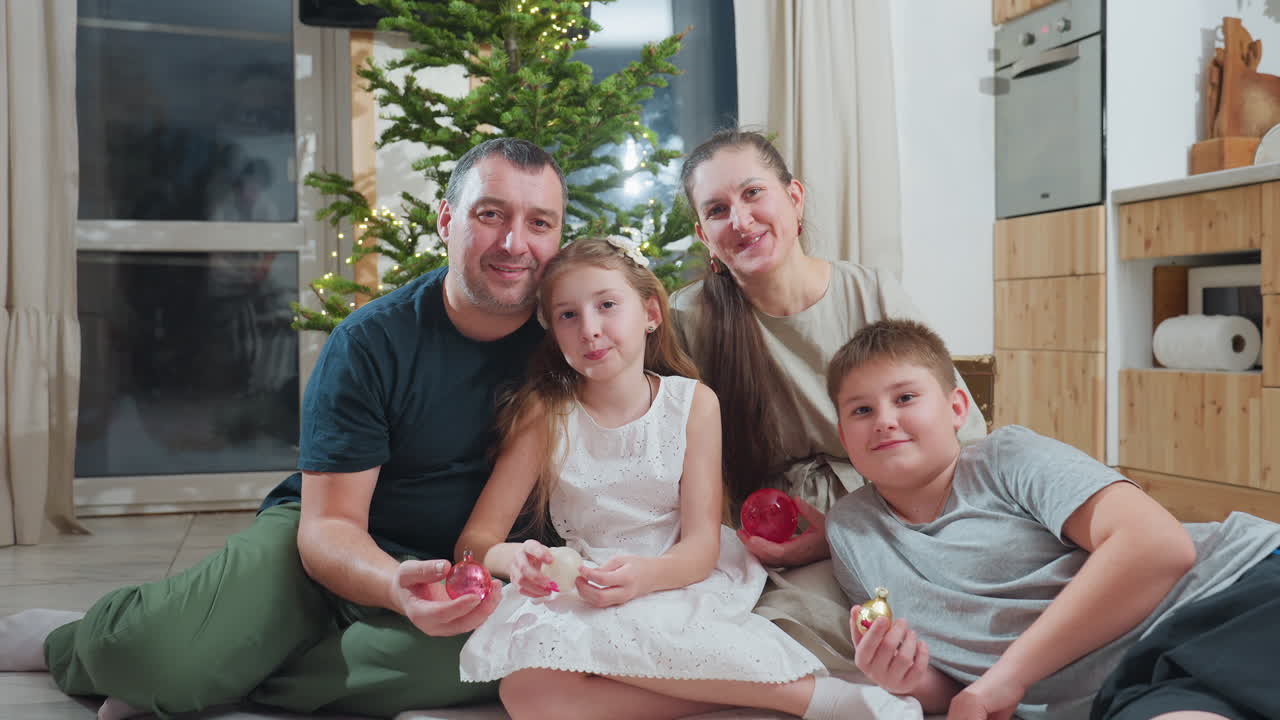 Close up view of cheerful family of four smiling warmly while posing for holiday photo together near beautifully decorated christmas tree in cozy modern home during festive winter celebration
