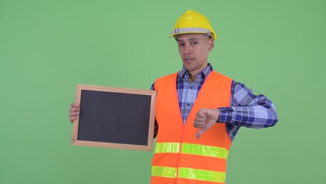 hombre multiétnico guapo trabajador de la construcción sosteniendo la pizarra y dando pulgares hacia abajo