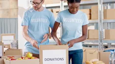 Hombre-Caucásico-Y-Joven-Voluntaria-Afroamericana-Empacando-Cajas-De-Donación-Con-Comida-En-Un-Almacén-De-Caridad