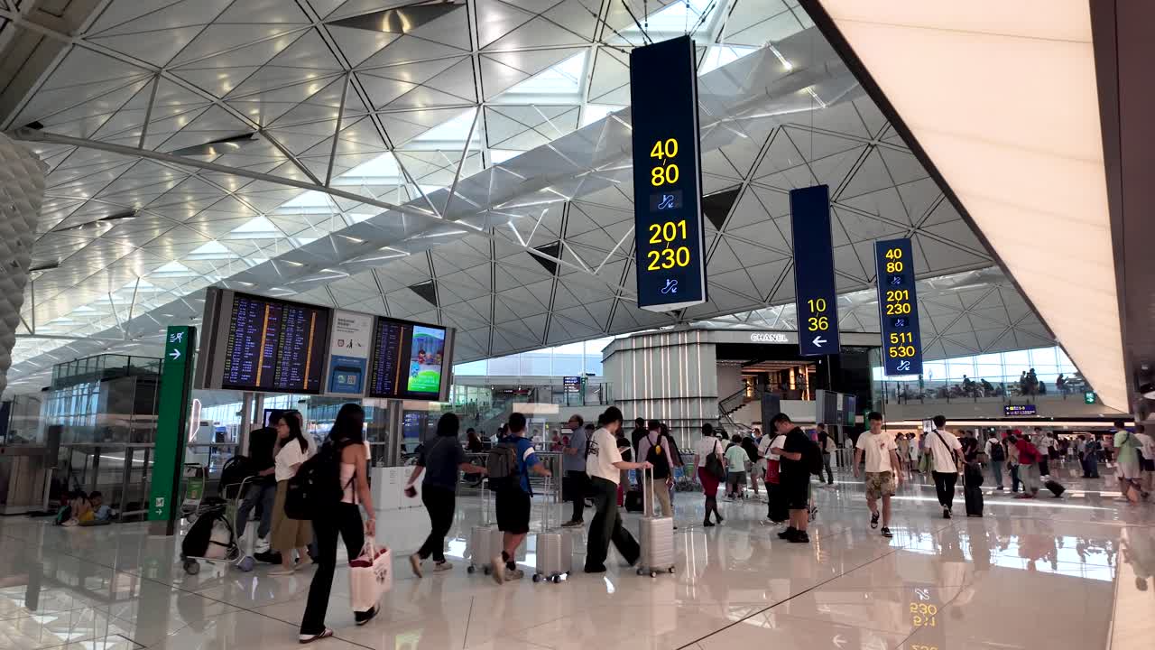 Travelers move through a busy airport terminal in Hong Kong, with visible gate information and departure boards.