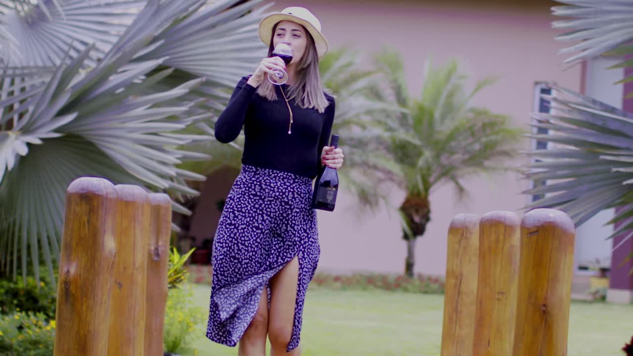 시골 와이너리를 걸으면서 레드 와인을 마시는 페도라를 입은 세련된 여성, 느린 동작