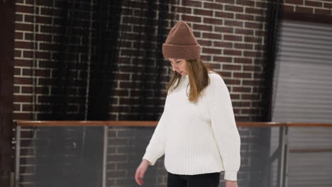 a close view of a woman leisurely skating on an ice rink, slowly moving while placing her hand on her head, she is wearing a white sweater and a brown beanie