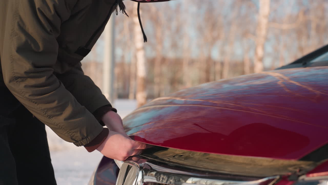 Teenage boy in brown hoodie and winter jacket attempts to open bonnet of red car on snowy ground while bending forward with focused expression, surrounded by bare trees and cold daylight atmosphere