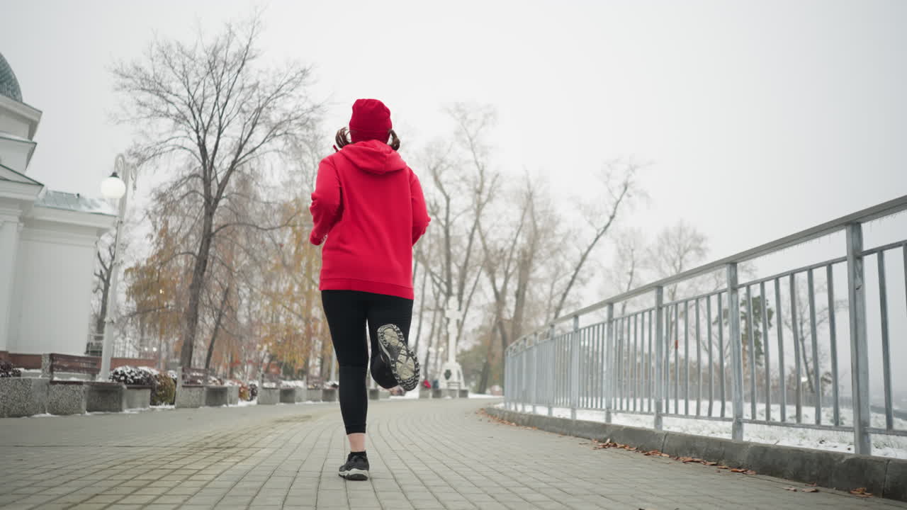 Back view of athlete jogging along snowy park pathway wearing red hoodie and black leggings, exercising near iron railing, surrounded by serene winter scenery, with white building by the side