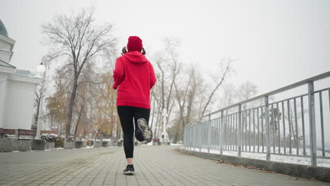 back view of athlete jogging along snowy park pathway wearing red hoodie and black leggings, exercising near iron railing, surrounded by serene winter scenery, with white building by the side