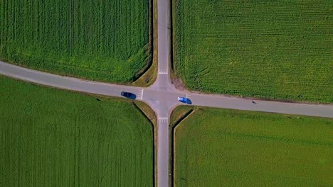 Vista-De-Pájaro-De-Un-Cruce-Simétrico-Perfecto-Con-Campos-Verdes-Y-2-Coches-Pasando