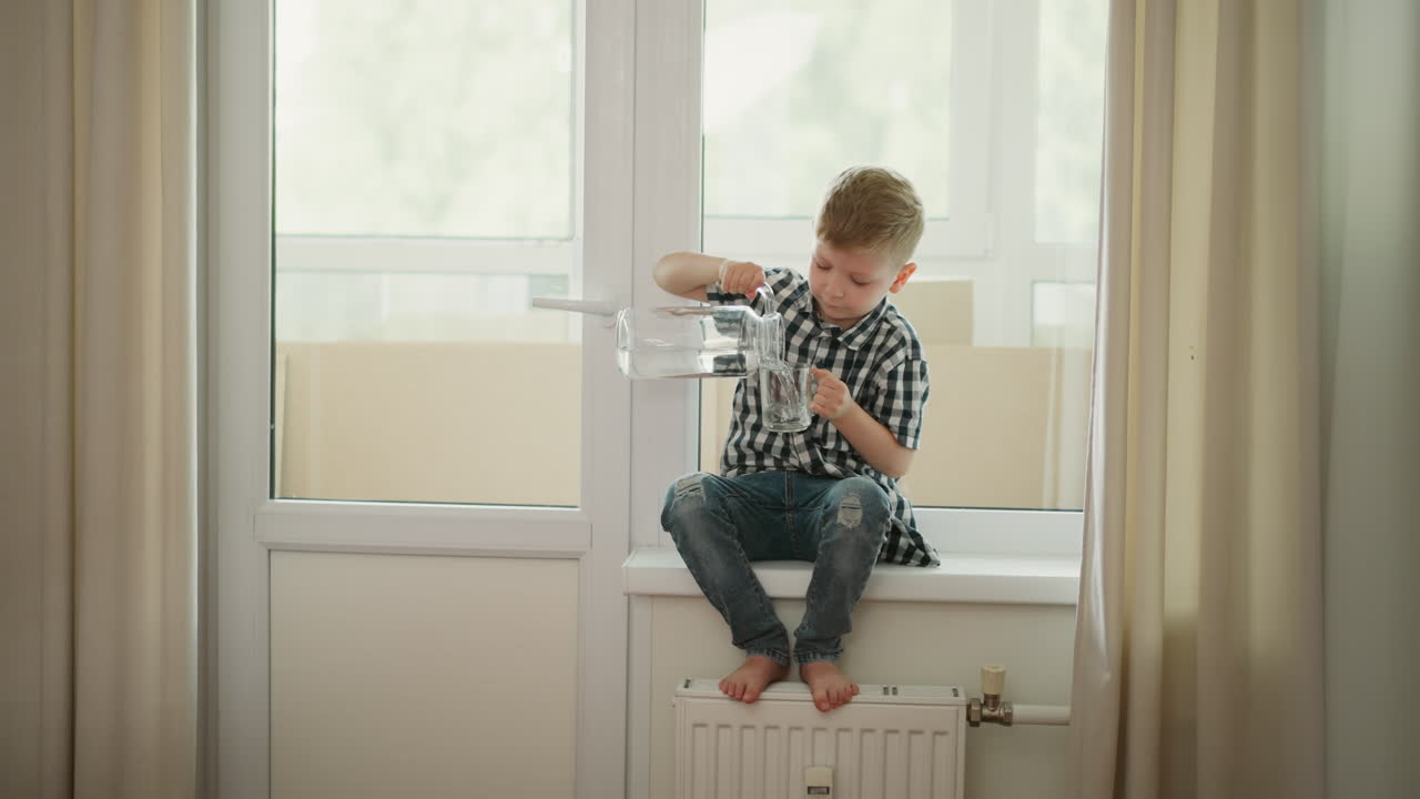 Boy pouring water from a jug into a glass on a window sill.