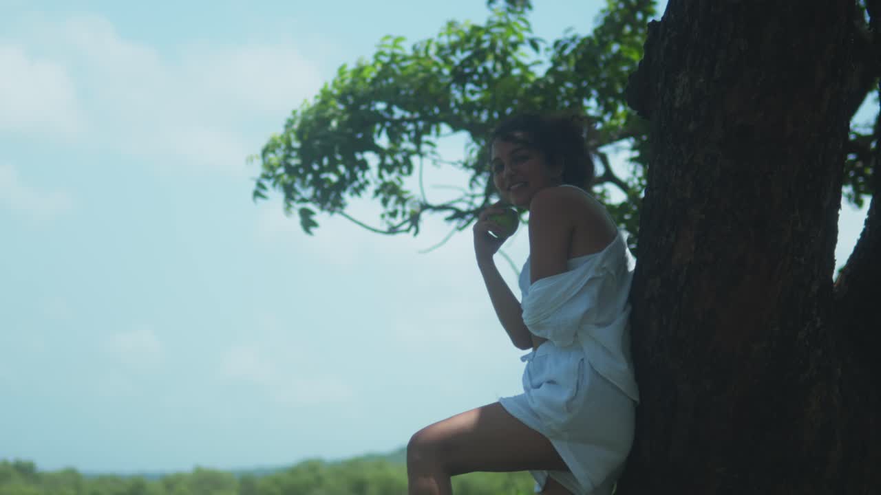 A woman in white stands with one knee raised, leaning on a tree and holding an apple. She smiles toward the camera under a bright sky