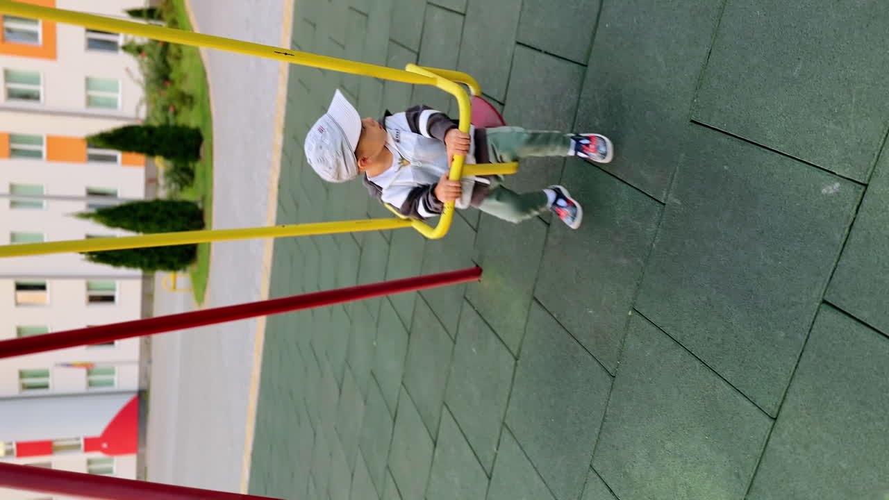 Calm baby boy sits on the swing outdoors. Toddler in a cap swinging and looking aside. Vertical view.