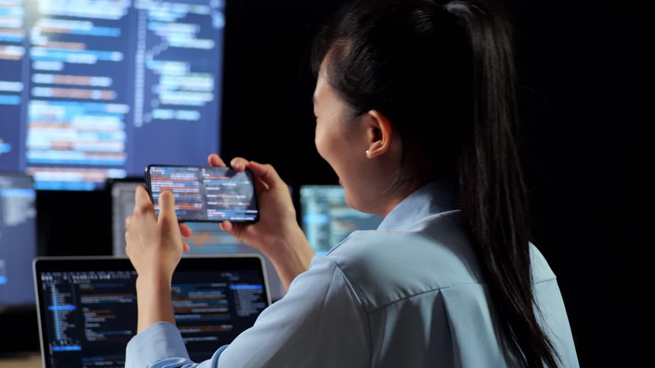 Close Up Back View Of Asian Female Programmer Looking At Smartphone Then Celebrating The Success While Writing Code By A Laptop Using Multiple Monitors Showing Data On Desktops In The Office