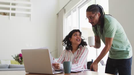 Smiling-biracial-woman-in-wheelchair-and-male-partner-doing-paperwork-using-laptop-in-living-room