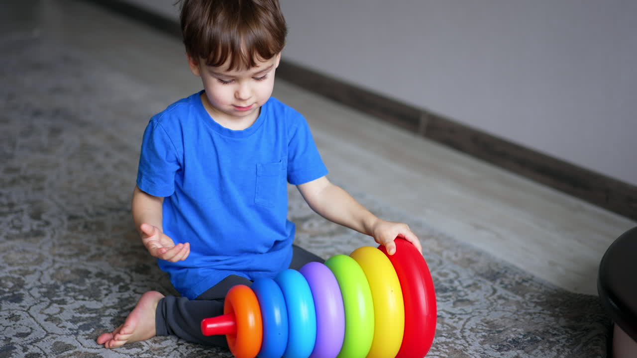 Calm Caucasian baby boy sitting on the carpet at home. Kid disassembles a toy pyramid and throws one ring aside.