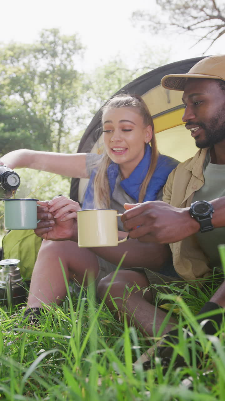 Vertical video of happy diverse couple camping, drinking tea and talking in park, slow motion