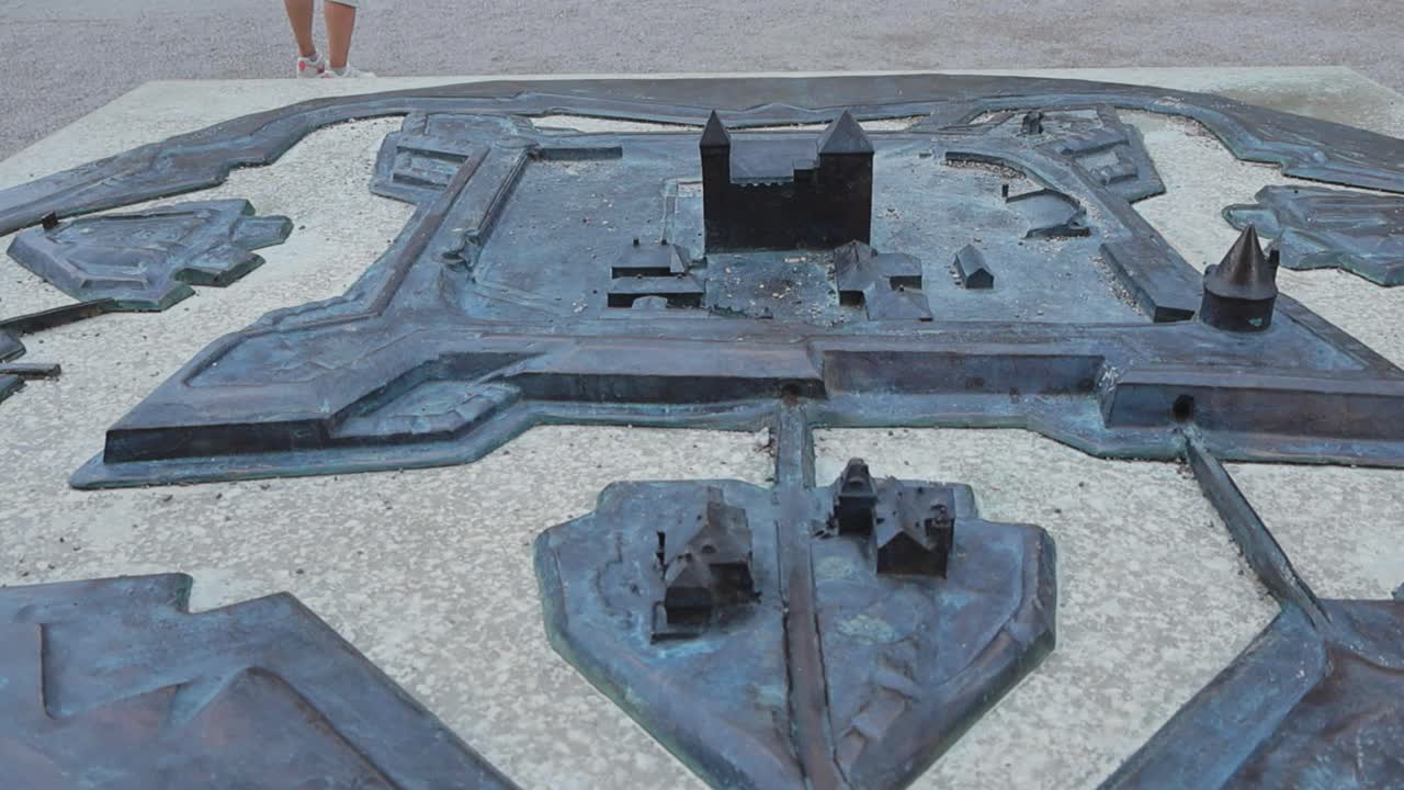 A metallic or bronze miniature model of Kuressaare castle and its passages, walls. The model is placed on a stone table outdoors during day time while it seems to be cloudy. Little castle visible.