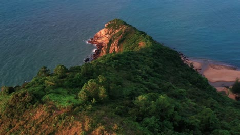 Aerial-rotation-looking-down-over-lush-tropical-Lantau-island-hilltop-slope-to-rippling-ocean-waves-coastline