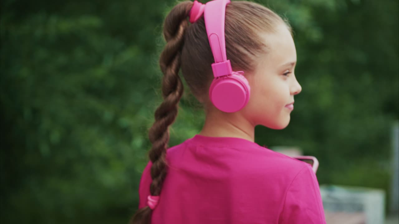 A young girl enjoys her music while wearing pink headphones and interacting with her smartphone, showcasing a moment of connectivity and joy amidst a vibrant green backdrop