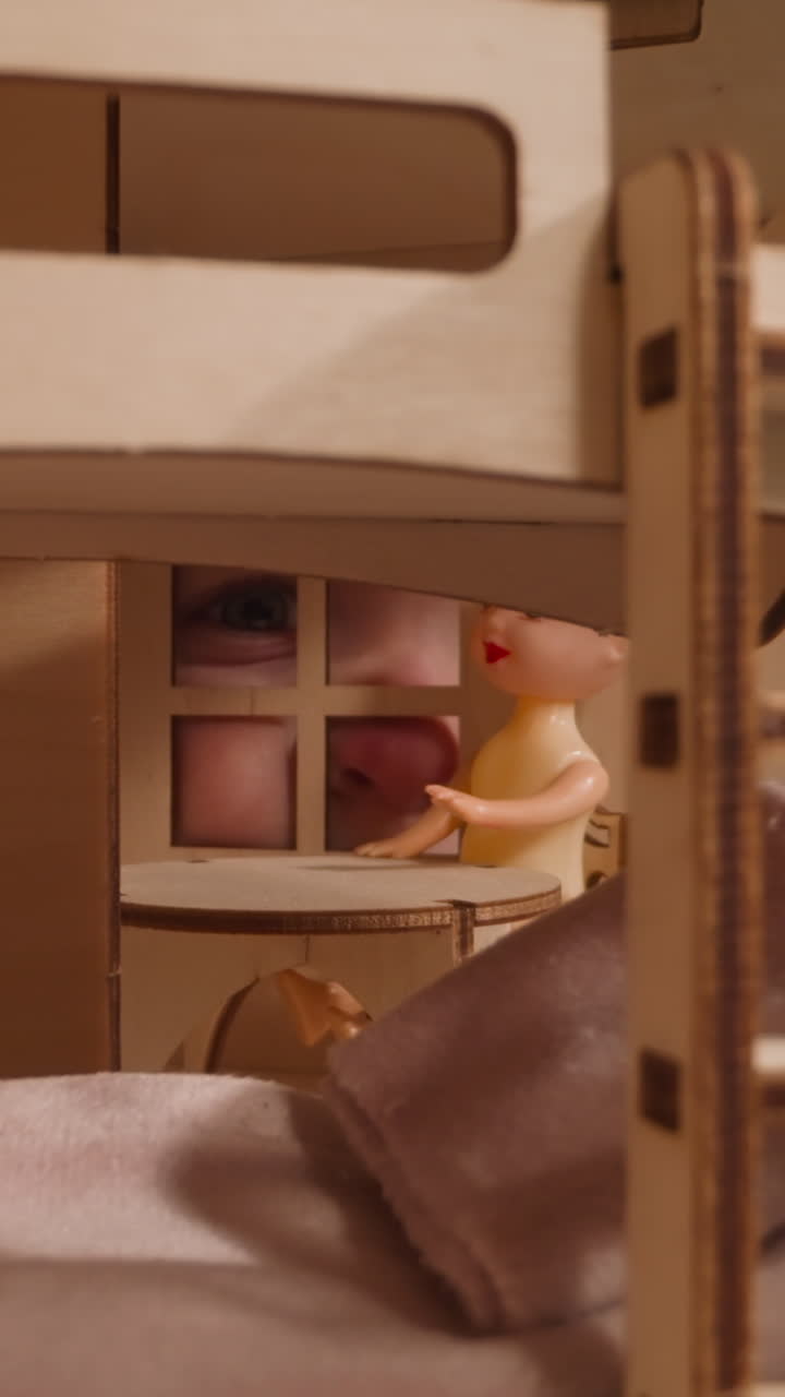 L'occhio azzurro di un bambino sbircia attraverso la finestra di una casa delle bambole fatta a mano con una piccola bambola seduta a un tavolo vicino all'armadio, vista ravvicinata con zoom indietro.