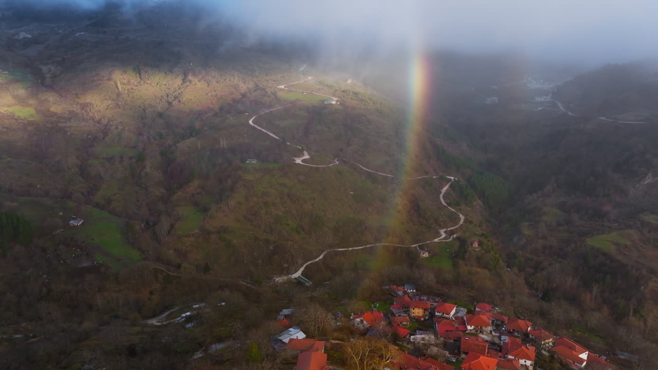 Flygvy i omloppsbana över Metsovo, Epirus, Grekland, med en dubbel regnbågsbåge ovanför bergdalen och hus med orange tak samt en slingrande väg