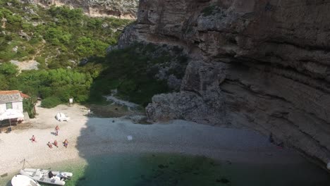 flying through a narrow gap between tall steep cliff toward a secluded beach with people sunbathing and boats moored in shallow clear water