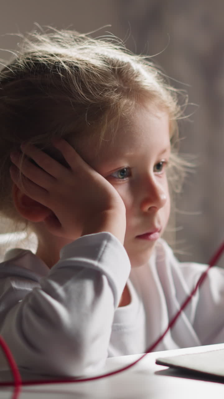 Bored little girl with braided hair watches educative video via tablet on holder sitting at table in light children room closeup slow motion