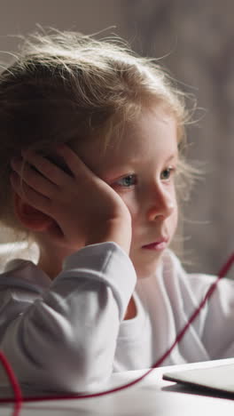 bored little girl with braided hair watches educative video via tablet on holder sitting at table in light children room closeup slow motion