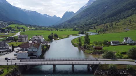 tourists traveling through olden city center over concrete bridge - closeup forward moving aerial - oldedalen mountain valley raging in background - norway