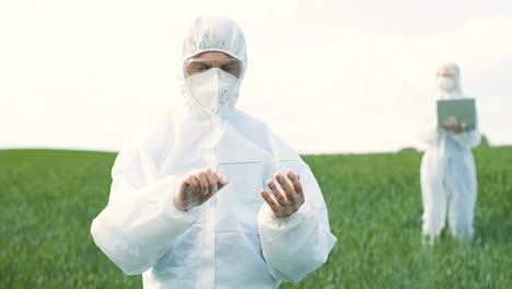 caucasian researcher man in protective suit and goggles tapping on glass in the green field