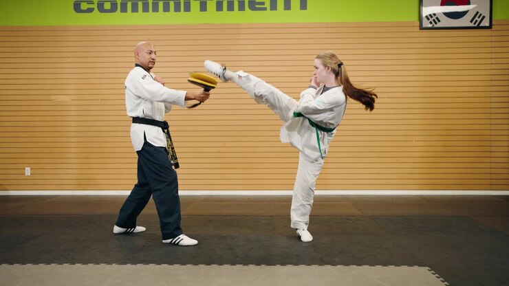 Wide Establishing Shot Young Female Combat Fighter Taekwondo Karate Martial Arts Student Slow Motion Powerful High Kick Into Pad with Black Belt Master Inside a Exercise Strength Gym Studio