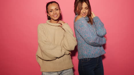 two women in sweaters posing against a pink background