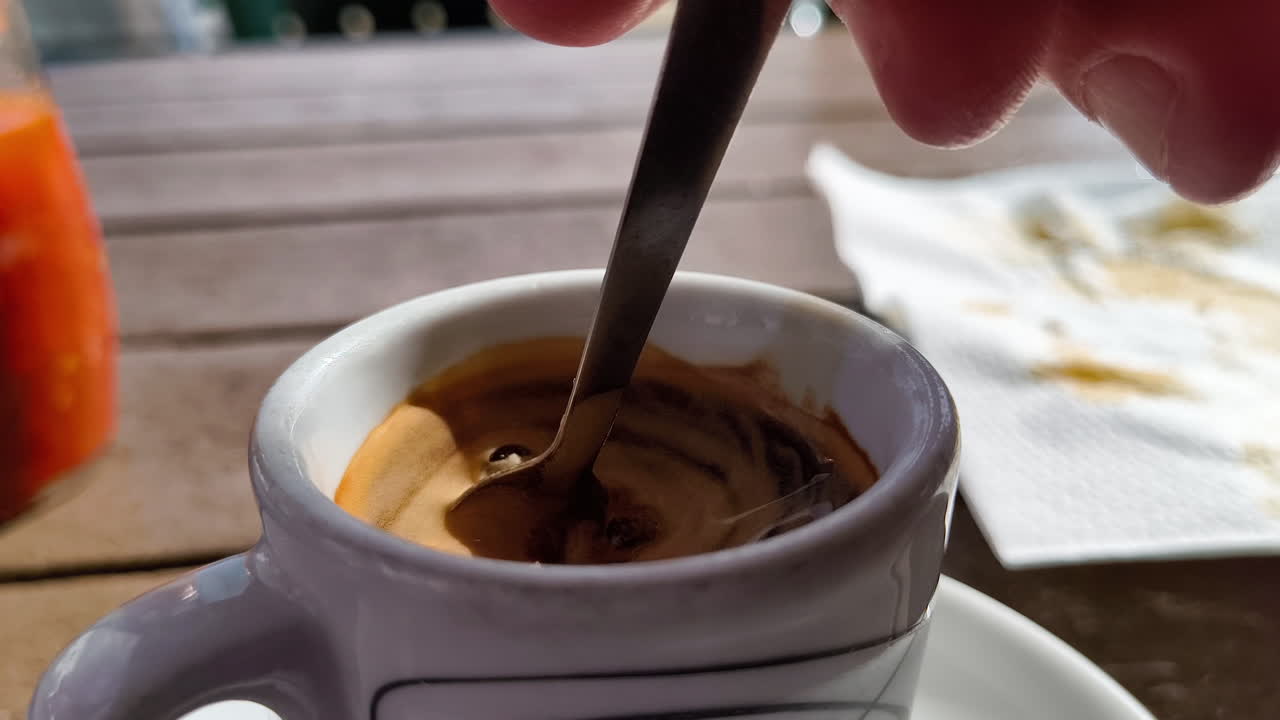 A close-up of a small white coffee cup with a dark espresso and a spoon stirring it. The crema on top of the coffee is thick and golden brown, and the liquid swirls around the spoon.
