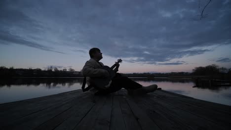 silhouette of musician in black playing guitar sitting on pier embankment on sunset