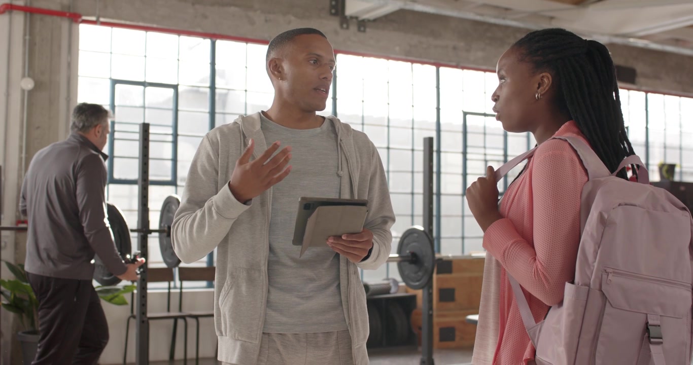 Trainer tapping tablet, gesturing, explaining gym plan to African American woman with pink backpack