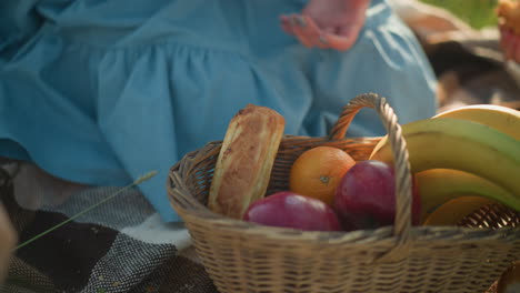 eine nahaufnahme einer picknickszene, in der ein mit früchten, äpfeln, bananen und einer orange gefüllter wickelkorb auf einem gefärbten schal sitzt. eine frau reicht ihre hand in den korb, um ein hot-dog-gebäck zu nehmen